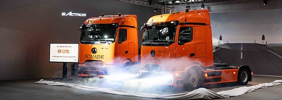 "Zwei orangefarbene Mercedes-Benz eActros-Lkw mit Schade-Logo w&auml;hrend einer Fahrzeug-Enth&uuml;llung mit Nebeleffekten.