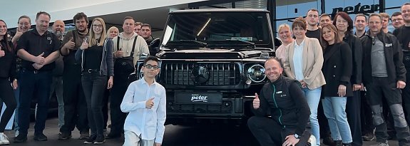 Jamil aus der TV-Show �The Voice Kids� steht mit einem Daumen-hoch-Zeichen vor einem schwarzen Mercedes-Gel&auml;ndewagen im Autohaus Mercedes Wittenberg, umgeben von Mitarbeiterinnen und Mitarbeitern des Autohauses.