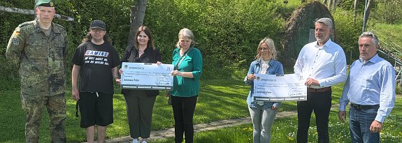 Gruppenfoto bei Spenden&uuml;bergabe zum 24-Stunden-Schwimmen: Sieben Personen stehen im Freien auf einer Wiese, darunter ein Soldat in Uniform, Teilnehmende in Freizeitkleidung und Vertreter des Autohauses Peter. Zwei gro&szlig;e Spendenschecks werden in die Kamera gehalten. Die Stimmung wirkt freundlich und gemeinschaftlich.