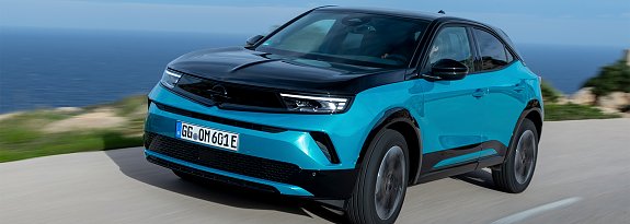 T&uuml;rkis-schwarzer Opel Mokka Electric f&auml;hrt auf einer K&uuml;stenstra&szlig;e, Meer und blauer Himmel im Hintergrund, Blick schr&auml;g von vorn.