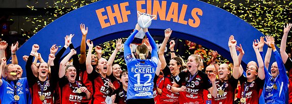 Das Bild zeigt ein Handballteam, das gerade den Sieg bei den EHF Finals feiert. In der Mitte hebt die Torh&uuml;terin mit der R&uuml;ckennummer 12 (Eckerle) den Pokal &uuml;ber den Kopf, w&auml;hrend gelbe Konfettistreifen durch die Luft wirbeln. Ihre Mitspielerinnen und das Trainerteam jubeln, klatschen und rei&szlig;en die Arme in die H&ouml;he. Alle tragen Medaillen und schwarze-rote Trikots, einige blaue Torwarttrikots. Im Hintergrund ist ein gro&szlig;er blauer Torbogen mit der Aufschrift EHF FINALS zu sehen.