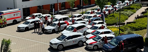 Gruppenfoto mit Fahrzeugflotte: Mehrere neue und bestehende Fahrzeuge des DRK Nordhausen, &uuml;berwiegend wei&szlig; mit rotem Kreuz, stehen in einem Innenhof. DRK-Mitarbeitende in roten Shirts posieren neben den Autos, im Vordergrund ein dunkelblauer Transporter.
