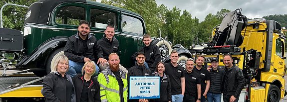 Gruppenfoto eines Teams des Autohaus Peter beim Festumzug in Sondershausen. Die Mitarbeitenden stehen vor einem klassischen Abschleppwagen des ADAC mit einem Oldtimer auf der Ladefl&auml;che. Alle tragen schwarze Autohaus-Peter-Bekleidung, einige lachen in die Kamera. Im Vordergrund wird ein Schild mit der Aufschrift Autohaus Peter GmbH gehalten. Im Hintergrund B&auml;ume und bew&ouml;lkter Himmel.