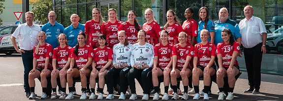 Gruppenfoto des Th&uuml;ringer Handball Clubs (THC) vor dem Autohaus Peter in Erfurt. In zwei Reihen posieren rund 20 Spielerinnen in roten Trikots gemeinsam mit Trainerteam und Partnern. Links und rechts au&szlig;en stehen Helmut Peter und Robert B&ouml;hm. Im Hintergrund mehrere Fahrzeuge mit Autohaus Peter-Branding. Das THC-Team l&auml;chelt geschlossen in die Kamera.