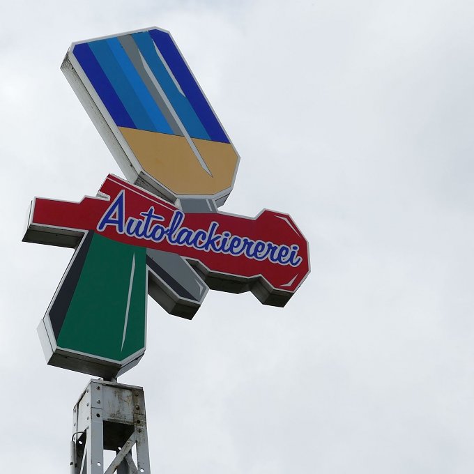 Werbeschild mit bunter Grafik in Form einer stilisierten Spr&uuml;hflasche und dem Schriftzug �Autolackiererei� in blauer und roter Schrift. Der Himmel ist grau und bew&ouml;lkt.