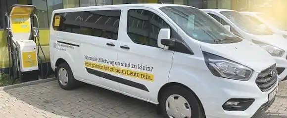 Wei&szlig;er Miet-Van mit ADAC-Beschriftung steht an einer Lades&auml;ule; seitlicher Schriftzug: Normale Mietwagen sind zu klein? Hier passen bis zu neun Leute rein.