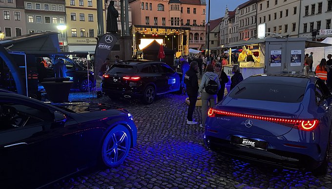 Das Bild zeigt die Eindr&uuml;cke der Aftershowparty. Die beleuchteten Mercedes-Benz Modelle strahlen in der Abendd&auml;mmerung mit Blick auf die B&uuml;hne der Veranstaltung. Zudem sind mehrere Interessierte G&auml;ste an unseren Fahrzeugen im Gespr&auml;ch.  