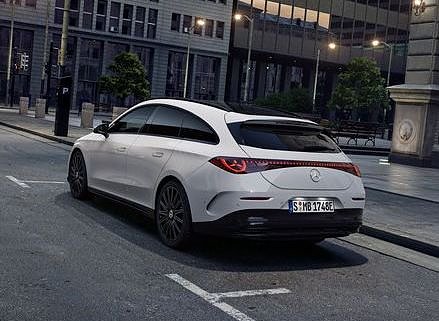 Das Bild zeigt einen wei&szlig;en Mercedes-Benz CLA Shooting Brake, der auf einer breiten, nahezu leeren Stadtstra&szlig;e steht. Moderne Geb&auml;ude umgeben die Szene. Der Wagen ist von hinten, seitlich zu sehen und vermittelt Dynamik sowie Eleganz.