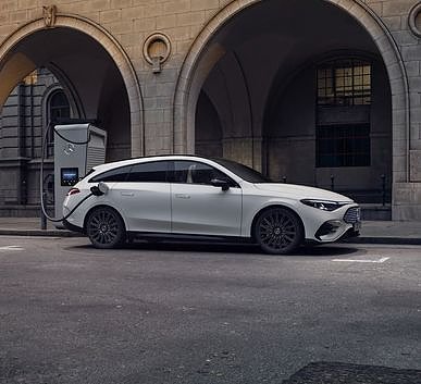Das Bild zeigt einen wei&szlig;en Mercedes-Benz CLA Shooting Brake neben einer Lades&auml;ule, der auf einer breiten, nahezu leeren Stadtstra&szlig;e steht. Moderne Geb&auml;ude umgeben die Szene, das Licht wirkt weich und fr&uuml;hmorgendlich. Der Wagen ist seitlich zu sehen und vermittelt Dynamik sowie Eleganz.