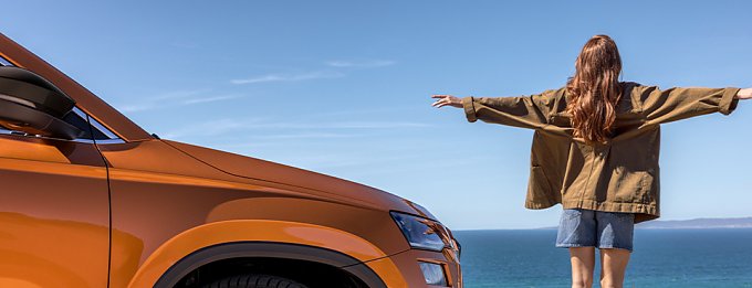 Frau steht mit ausgebreiteten Armen am Meer neben orangefarbenem SUV.
